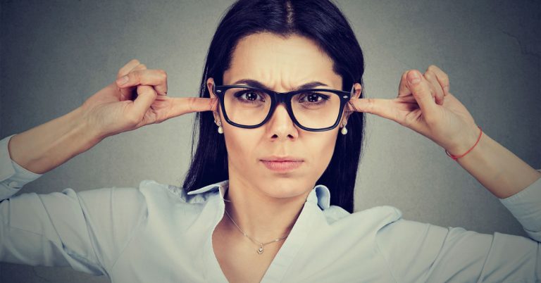 A white woman with long dark hair wearing black glasses as she puts her fingers in her ears. Arrogance can make us deaf to anyone else's opinions.